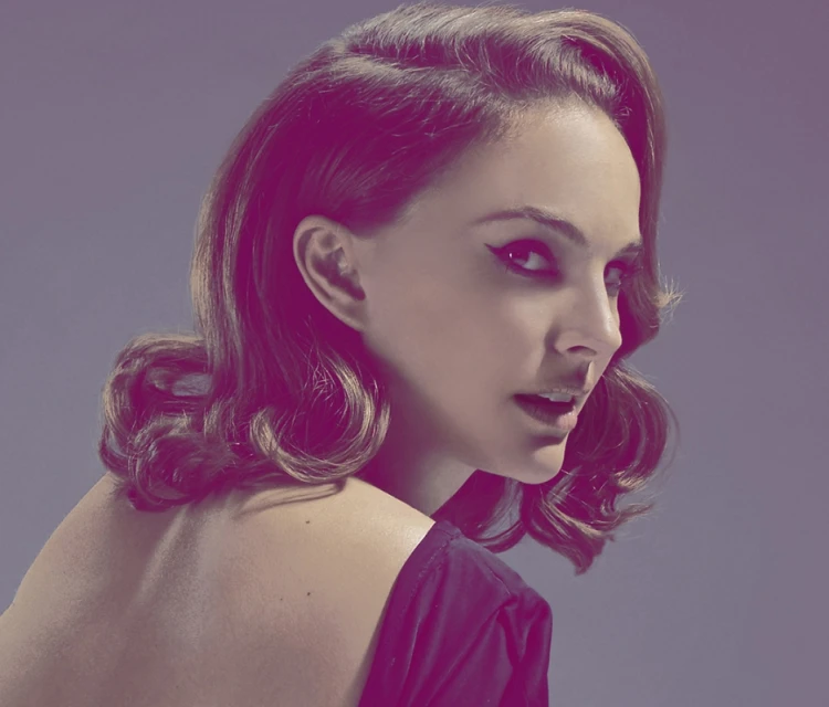 Glamorous headshot of a young adult woman with retro-inspired hair and dramatic eyeliner, posing against a solid lavender background.