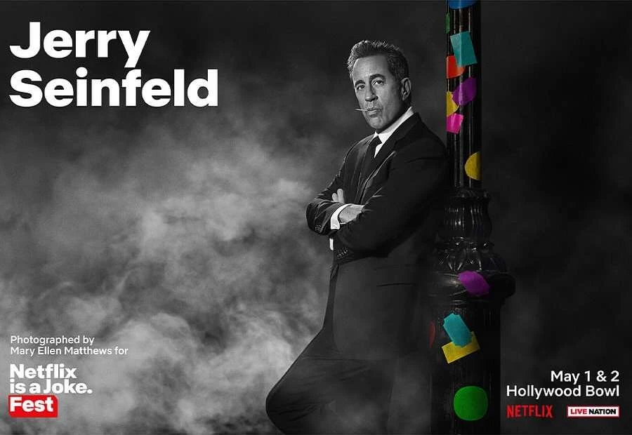 Black-and-white portrait of Jerry Seinfeld leaning against a black lamppost with colorful accents, set against a smoky backdrop.