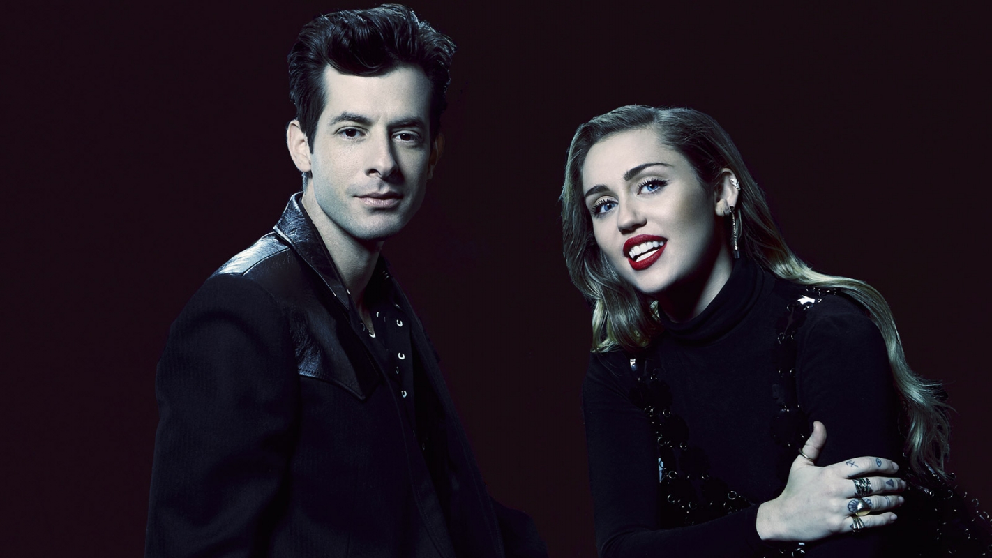 Mark Ronson and Miley Cyrus pose in dark studio. Ronson wears black leather jacket, Cyrus a black turtleneck, both with neutral expressions.