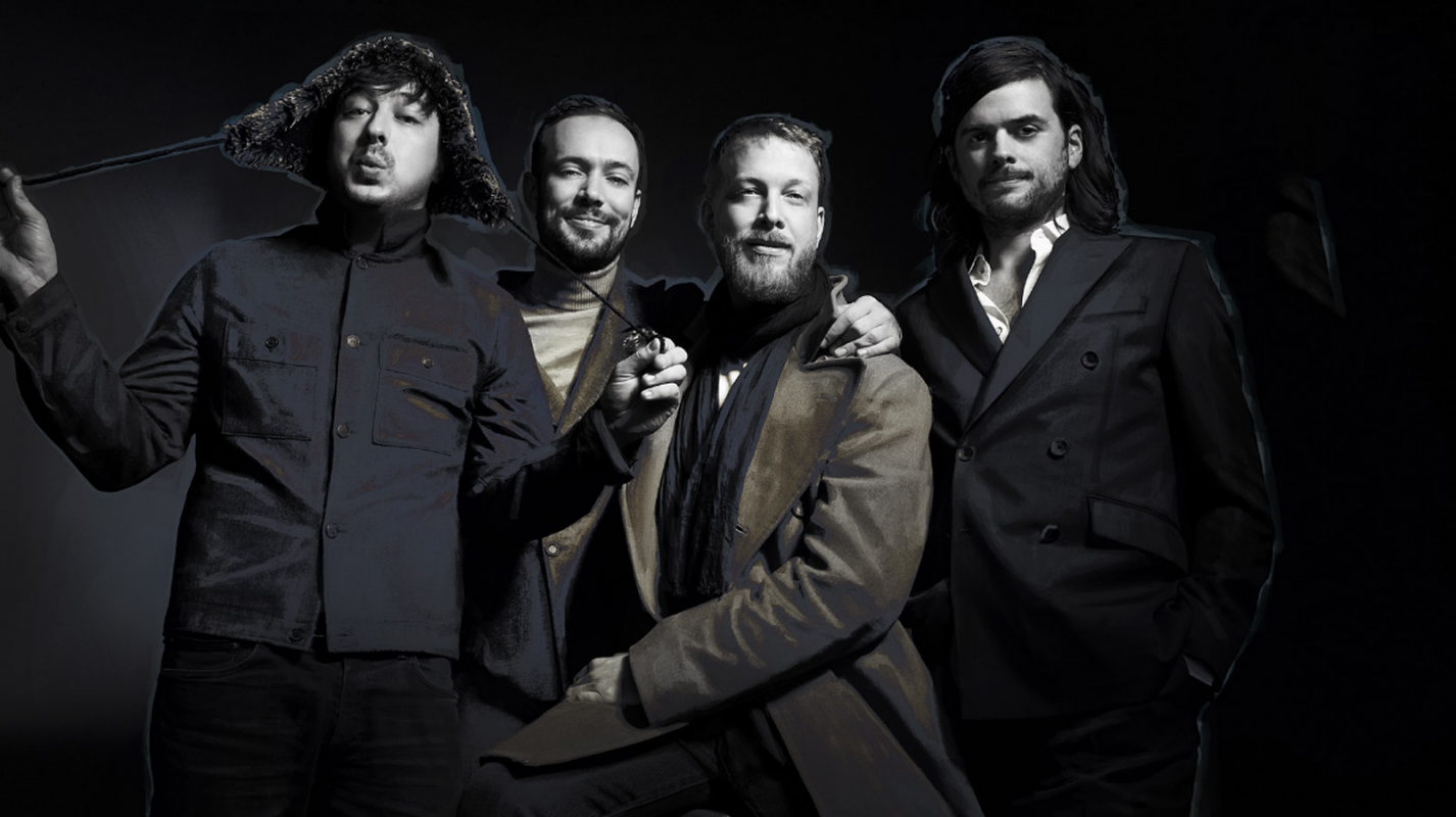 Group studio shot of four adult men in formal wear with dark background, one wearing a fur hat and holding an object with a long handle.