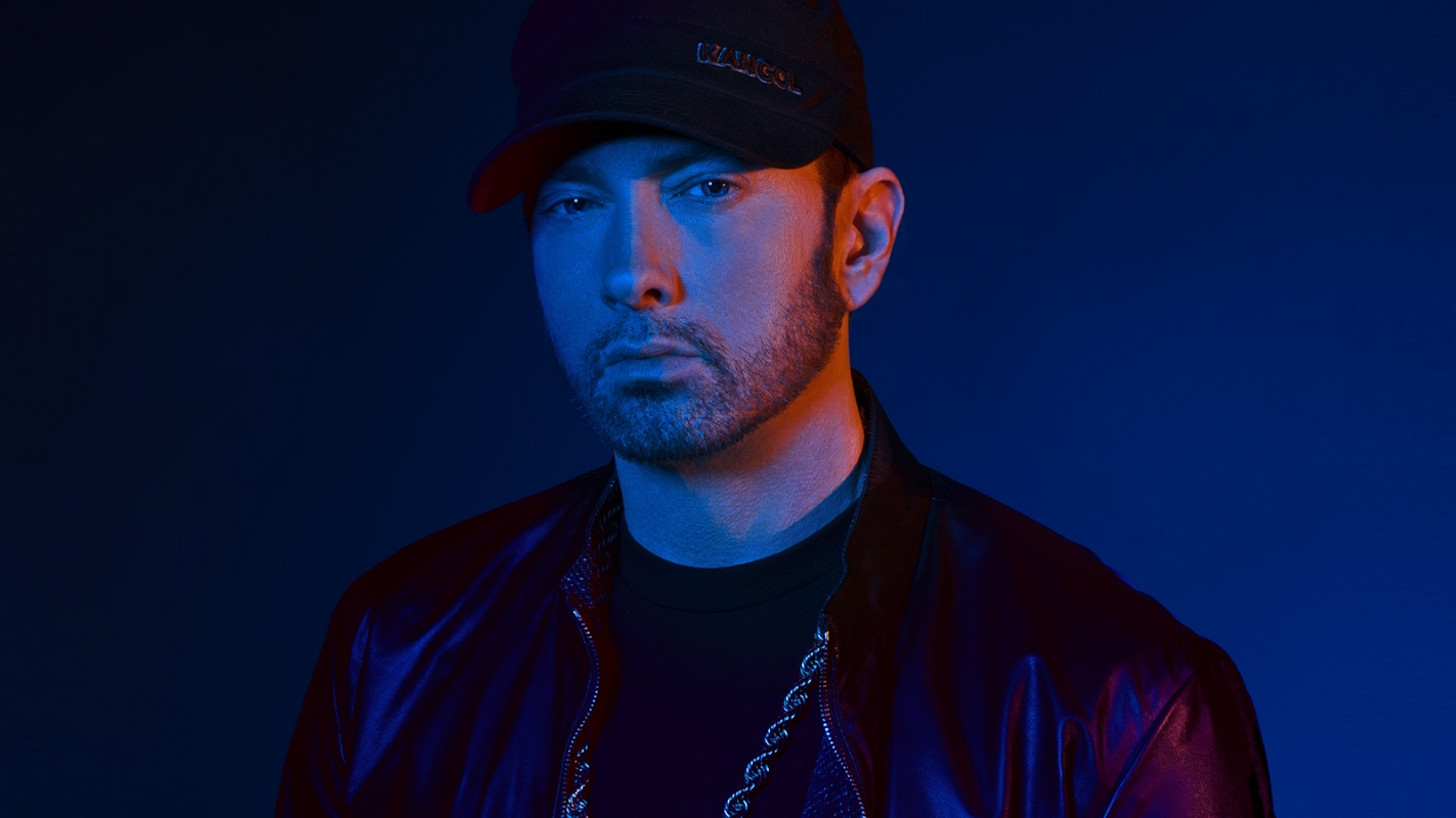 Portrait of Eminem wearing a black baseball cap and jacket, highlighted with blue and red lighting against a dark blue background.