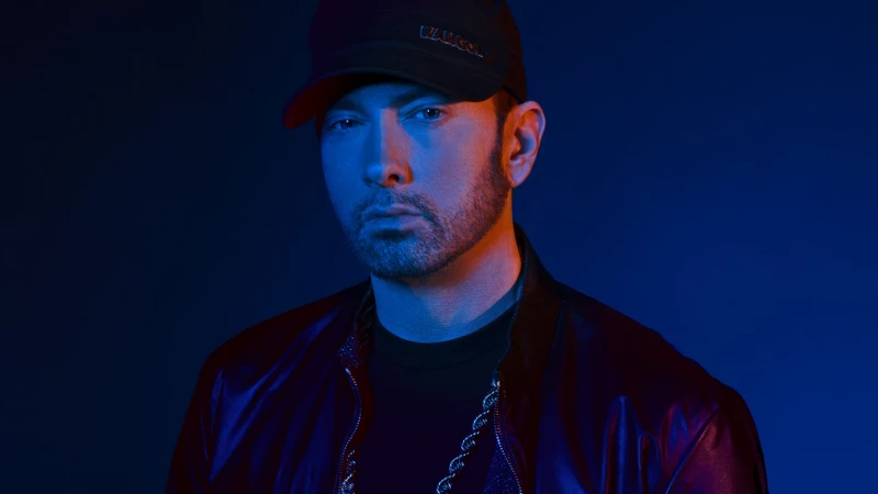 Eminem wearing a black cap and jacket, illuminated by moody blue and red lighting against a dark blue background.