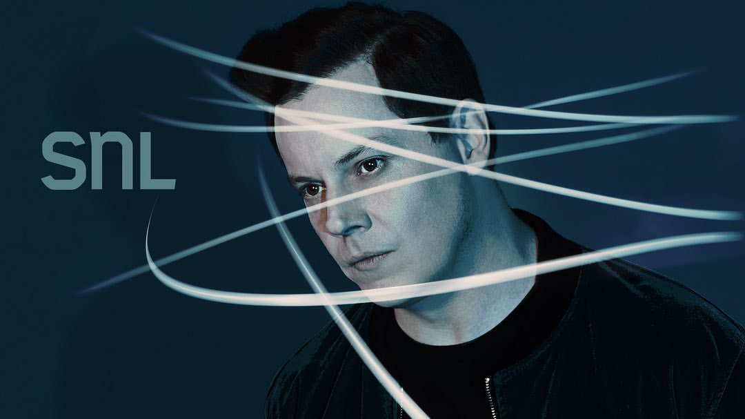 Portrait of an adult male with dark hair wearing a black bomber jacket with light lines digitally added over a dark blue background.