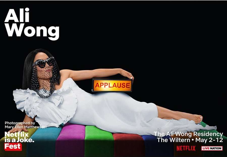 Promotional image of Ali Wong lying on a striped surface, wearing a white gown, sunglasses, and veil, holding an 'Applause' sign.