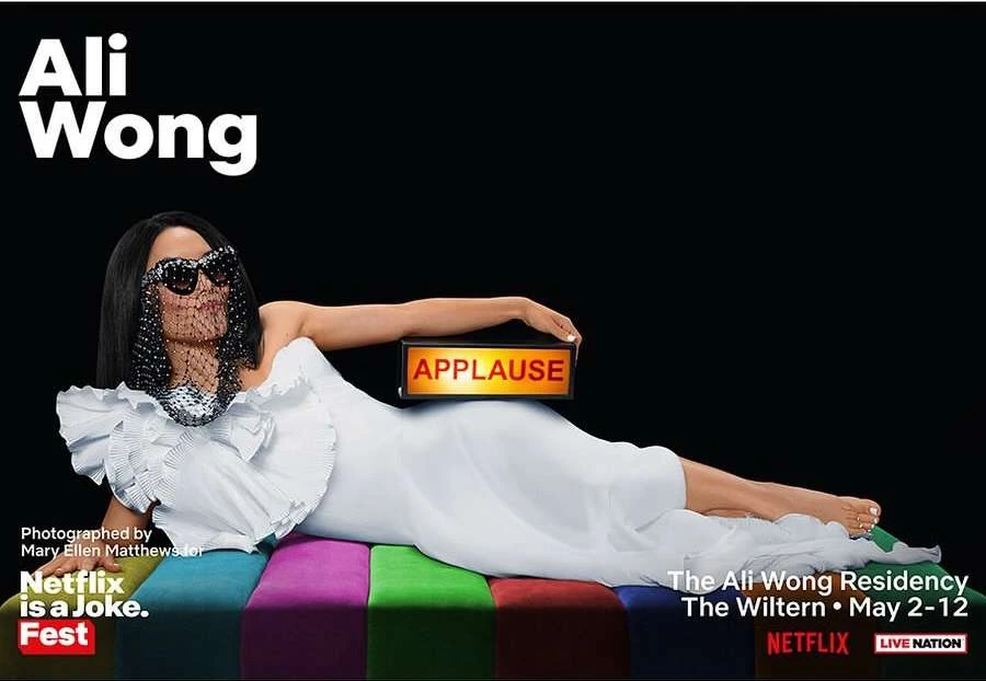 Promotional image of Ali Wong lying on a striped surface, wearing a white gown, sunglasses, and veil, holding an 'Applause' sign.