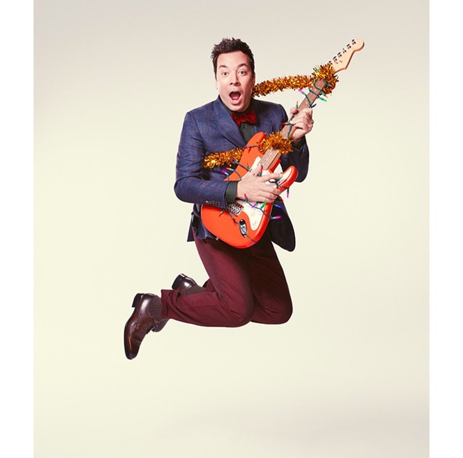 Jimmy Fallon leaps mid-air holding a decorated electric guitar. He wears a navy blazer, maroon pants, and dress shoes against a plain backdrop.
