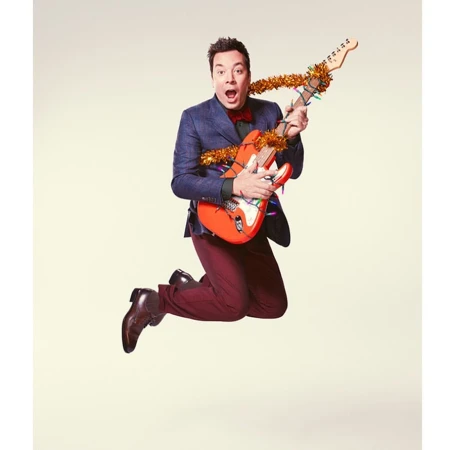 Jimmy Fallon leaps mid-air holding a decorated electric guitar. He wears a navy blazer, maroon pants, and dress shoes against a plain backdrop.