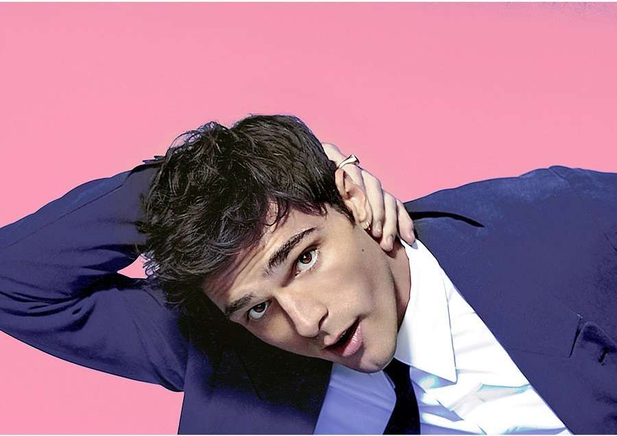 Headshot of a brunette adult male in a dark suit with a light collared shirt and tie, striking a pose against a pink background.