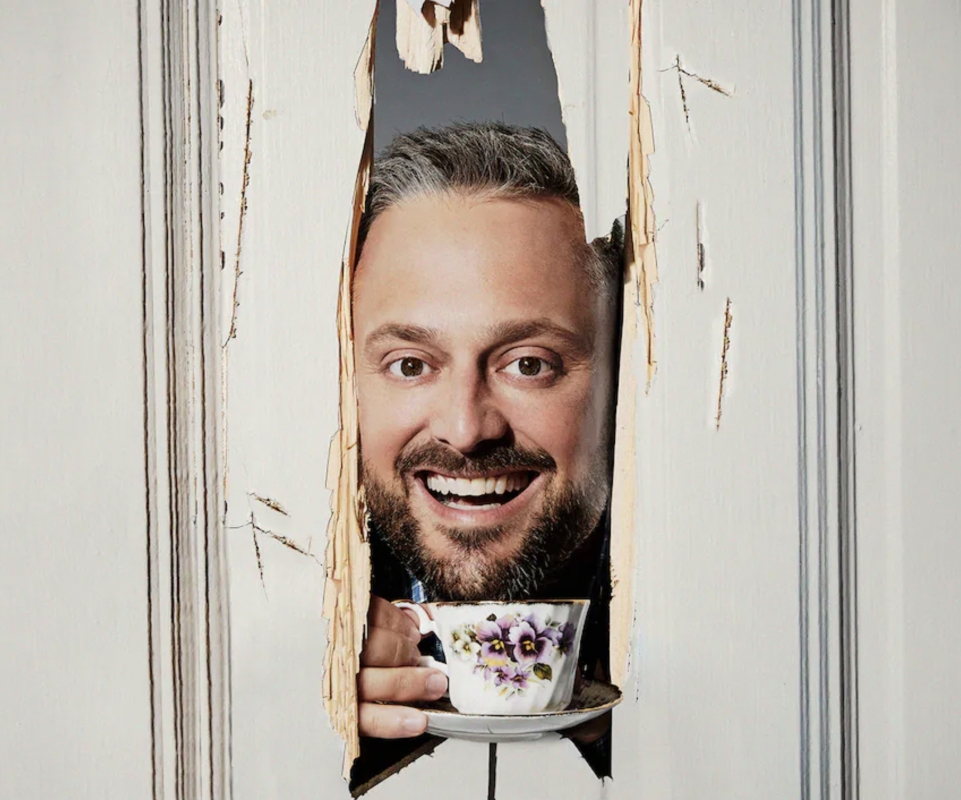 Bearded man smiles, peering through splintered white door, holding a floral teacup. Indoor setting with natural lighting.