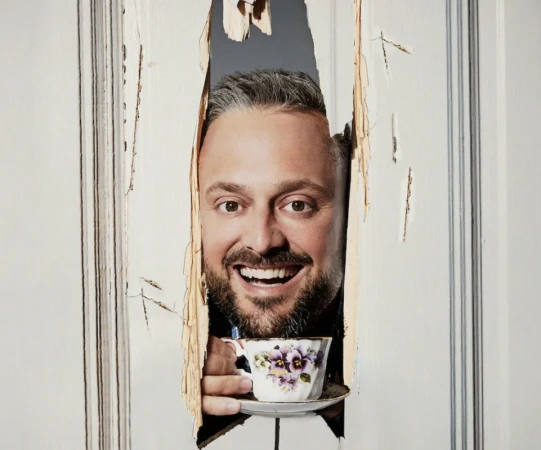 Bearded man smiles, peering through splintered white door, holding a floral teacup. Indoor setting with natural lighting.