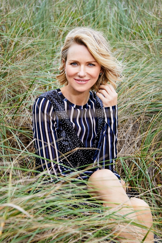 Blonde woman in a striped dress poses in tall green grass, smiling at the camera. Natural outdoor setting, medium shot.