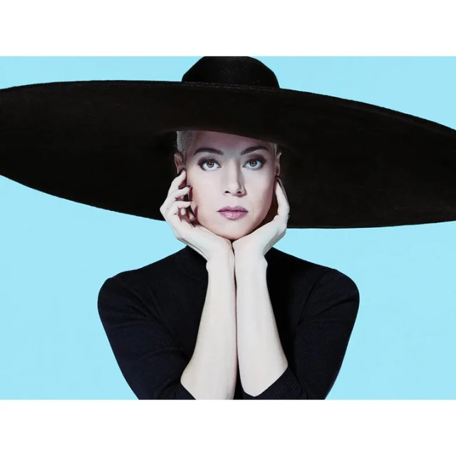 Woman wearing a wide-brimmed hat and turtleneck sweater against a light blue backdrop, hands gently framing her face in a fashion pose.