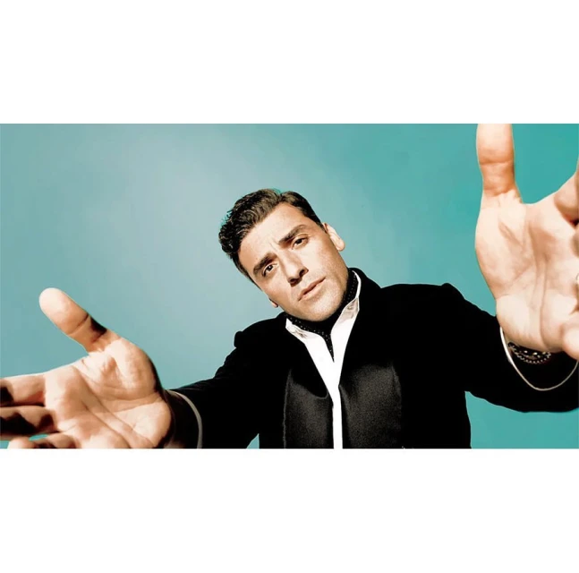 Portrait of Oscar Isaac in a black suit with hands outstretched towards the camera, set against a plain aqua background.