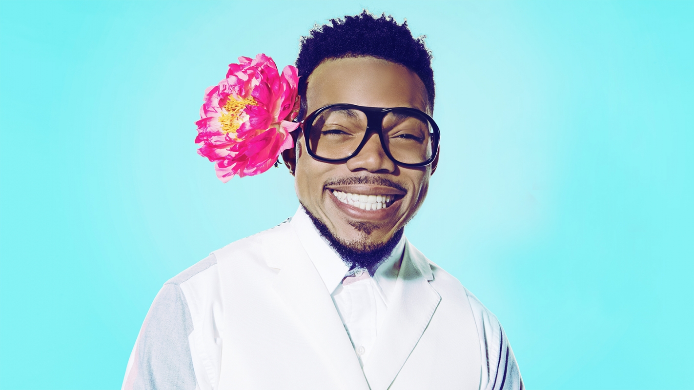 Close-up studio shot of an adult man smiling with glasses and a flower over his ear, set against a bright turquoise backdrop.