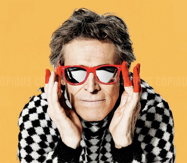 Portrait of an adult male with gray hair wearing red sunglasses, touching his face with paint-covered fingers, against a yellow backdrop.