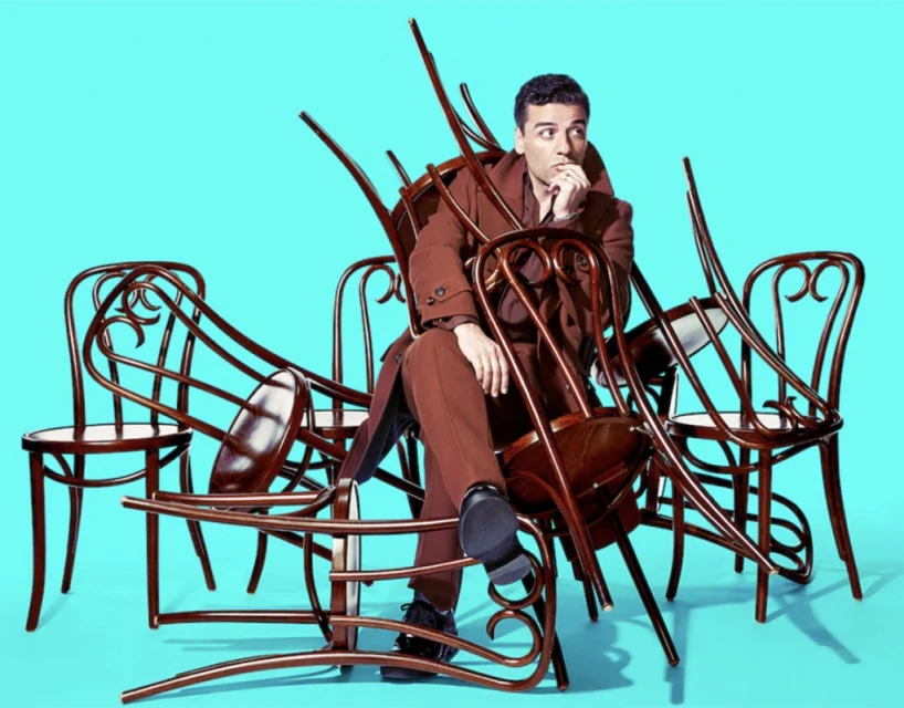 Adult male in brown coat sits surrounded by stacked brown bentwood chairs against a turquoise background in a medium studio shot.