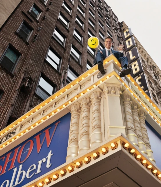 Man in suit sitting on ledge of theatre building with smiling yellow balloon. Urban shot with architecture and sign. 