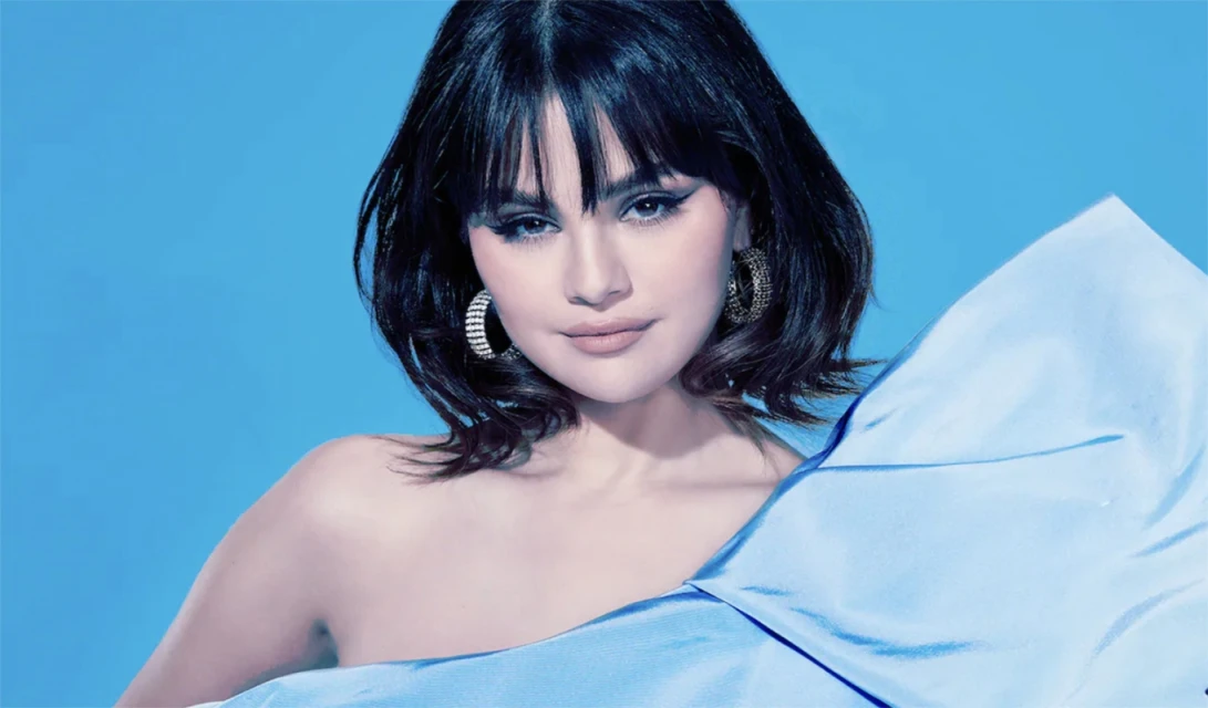 Close-up portrait of a brunette woman with bangs, wearing hoop earrings and a blue silk wrap, set against a solid blue background.