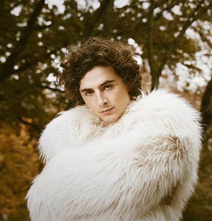 A portrait of a young man with curly brown hair wearing a white fur coat, posing outdoors with a neutral expression.