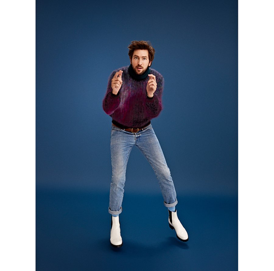 Man wearing a textured purple sweater and light blue jeans posing against a solid blue background