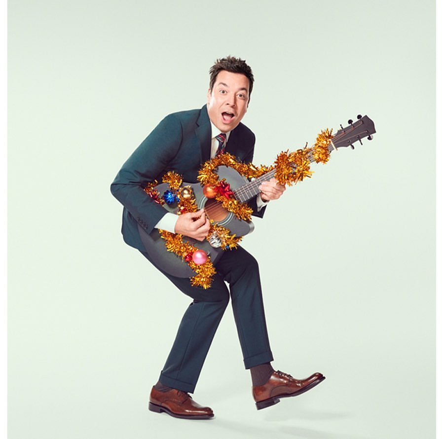 Enthusiastic man in suit plays guitar with Christmas garland and ornaments, posing in front of a light background in a studio.