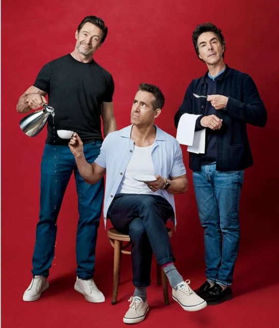 Three adult men, Hugh Jackman, Ryan Reynolds, and unknown male, pose in front of a red background, holding a teapot, cup, spoon, and towel.
