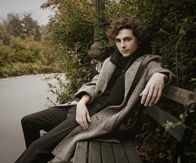 A male model with curly hair sits on a park bench wearing a herringbone coat and a black scarf in an outdoor portrait.