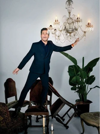 A man in a dark suit balancing on a chair among scattered antique furniture and a hanging crystal chandelier.