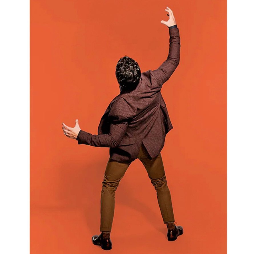 Adult male in burgundy blazer and brown trousers stands with back to camera on orange background with hands raised overhead in studio shot.
