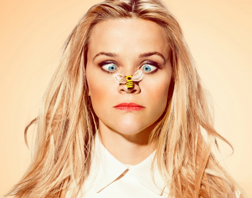 Blonde woman with blue eyes makes a silly face, cross-eyed with a small bee figurine on her nose in a studio portrait. 