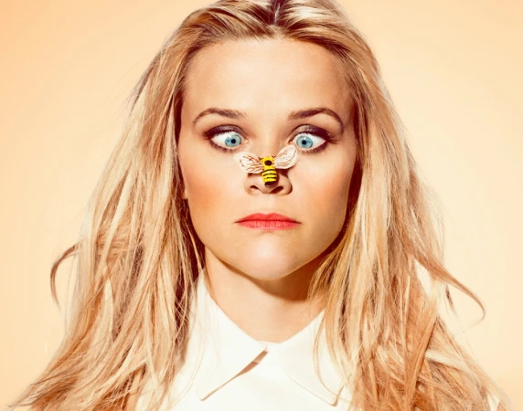 Blonde woman with blue eyes makes a silly face, cross-eyed with a small bee figurine on her nose in a studio portrait. 