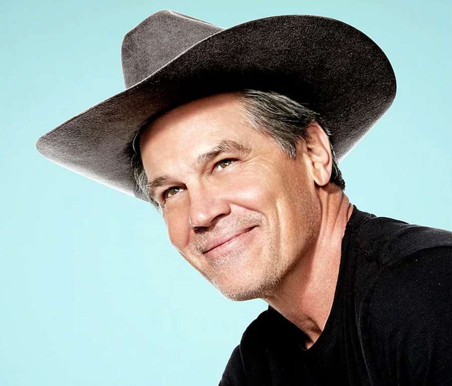 Portrait of mature adult male in black shirt wearing a grey cowboy hat against a light blue backdrop, smiling and looking upward.