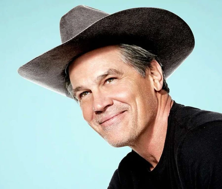 Portrait of mature adult male in black shirt wearing a grey cowboy hat against a light blue backdrop, smiling and looking upward.