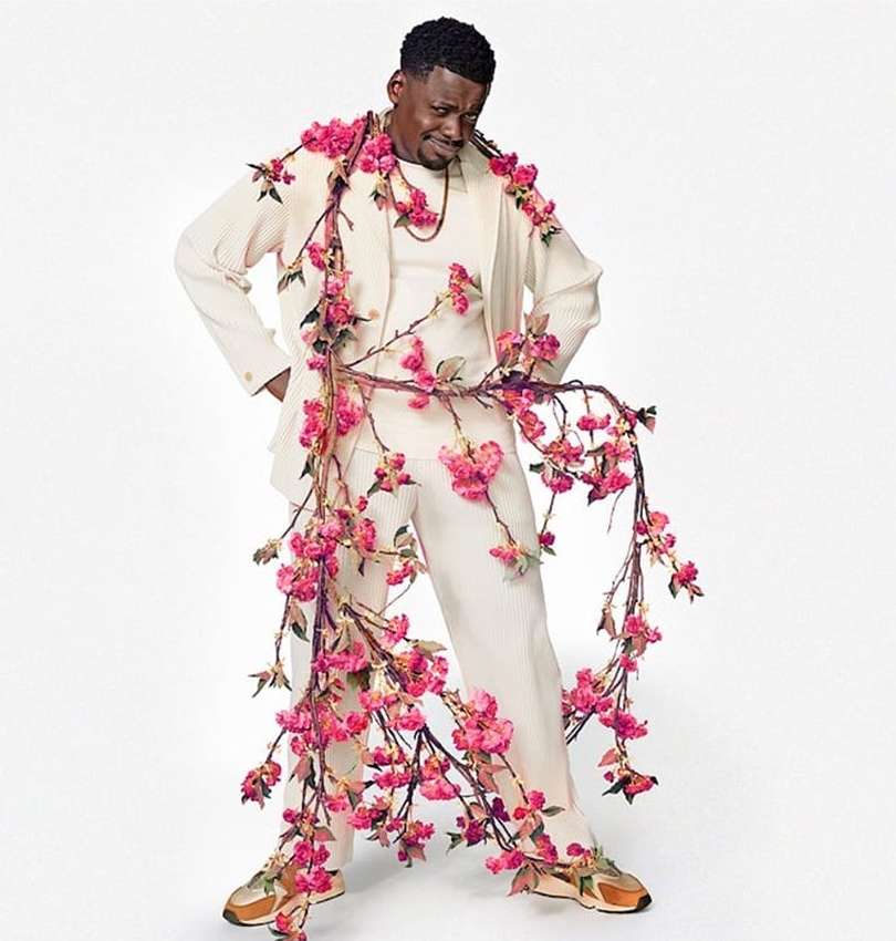 Adult male wearing a cream suit adorned with pink flowers posing in a photo studio with neutral background and looking away from the camera.