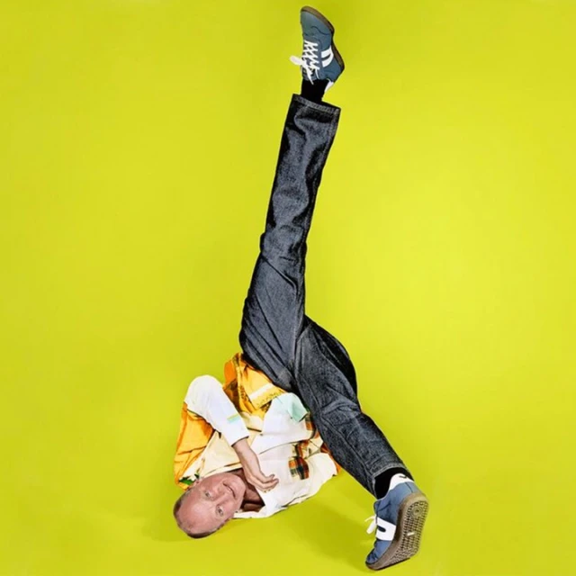 Adult male in a headstand pose against a vibrant yellow backdrop, wearing jeans, sneakers, and a plaid shirt, showcasing fitness and fashion.