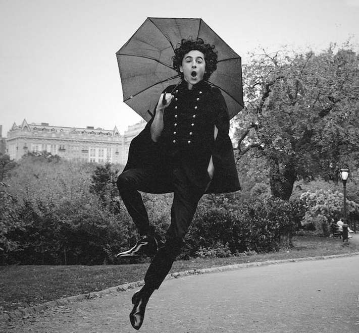 Black and white photo of a person in vintage clothing mid-air, holding an umbrella, with a building and trees in the background.