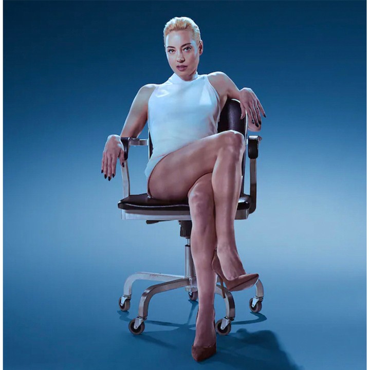 Woman in white top and brown heels sits on an office chair in a modern photo studio with a blue gradient background and overhead lighting.