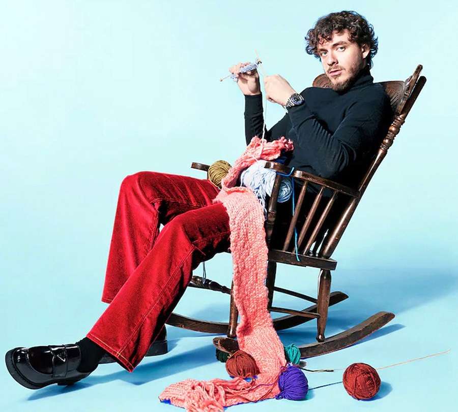 Jack Harlow in a wooden rocking chair, knitting with yarn and needles against a light blue backdrop in a celebrity and entertainment themed portrait.