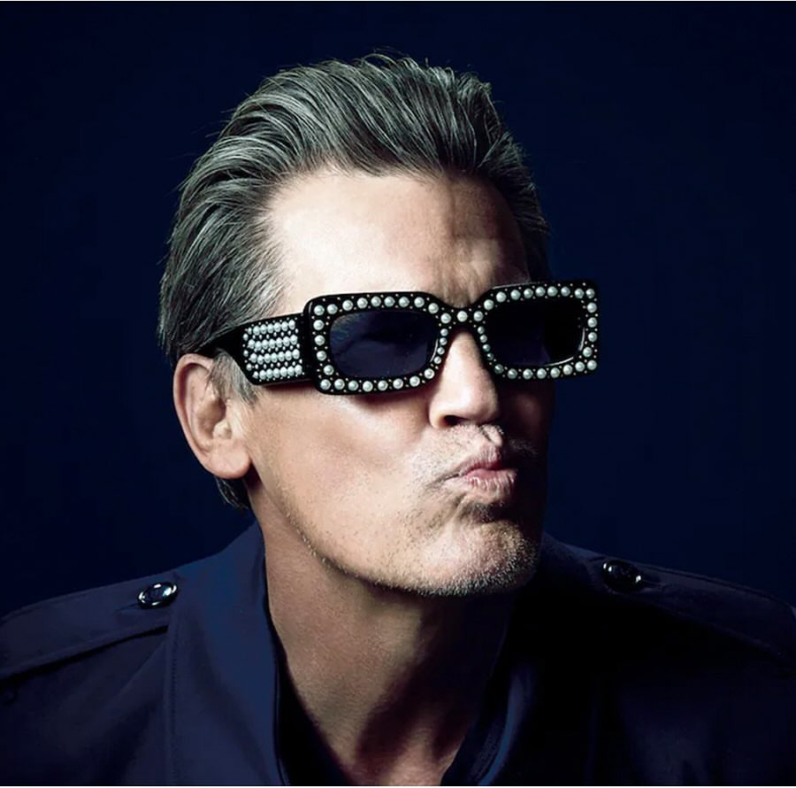 Man with gray hair poses wearing sunglasses with pearl-like decorations against a dark background in a chest-up studio shot.