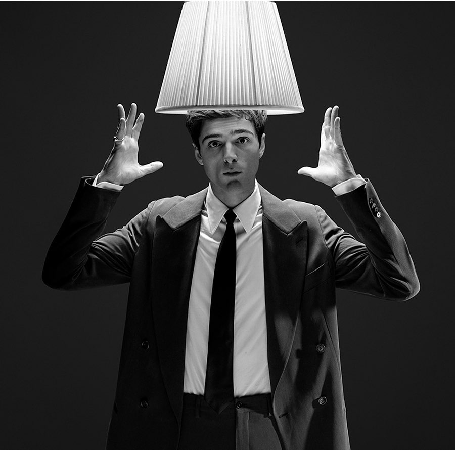 Black and white portrait of a man in a suit with a lampshade above his head, holding his hands up on either side of it.