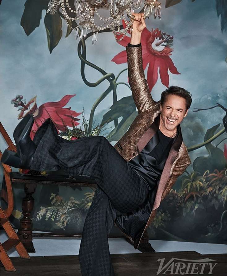 Robert Downey Jr. poses indoors with an arm raised, wearing a black top, patterned black pants, and a glittery gold jacket, seated on furniture.