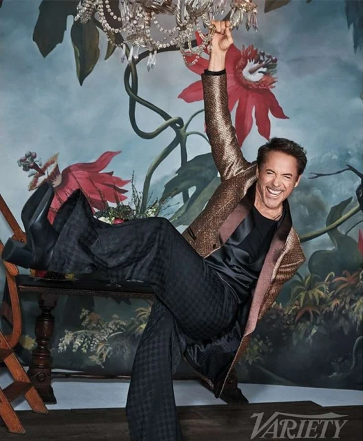Robert Downey Jr. poses indoors with an arm raised, wearing a black top, patterned black pants, and a glittery gold jacket, seated on furniture.
