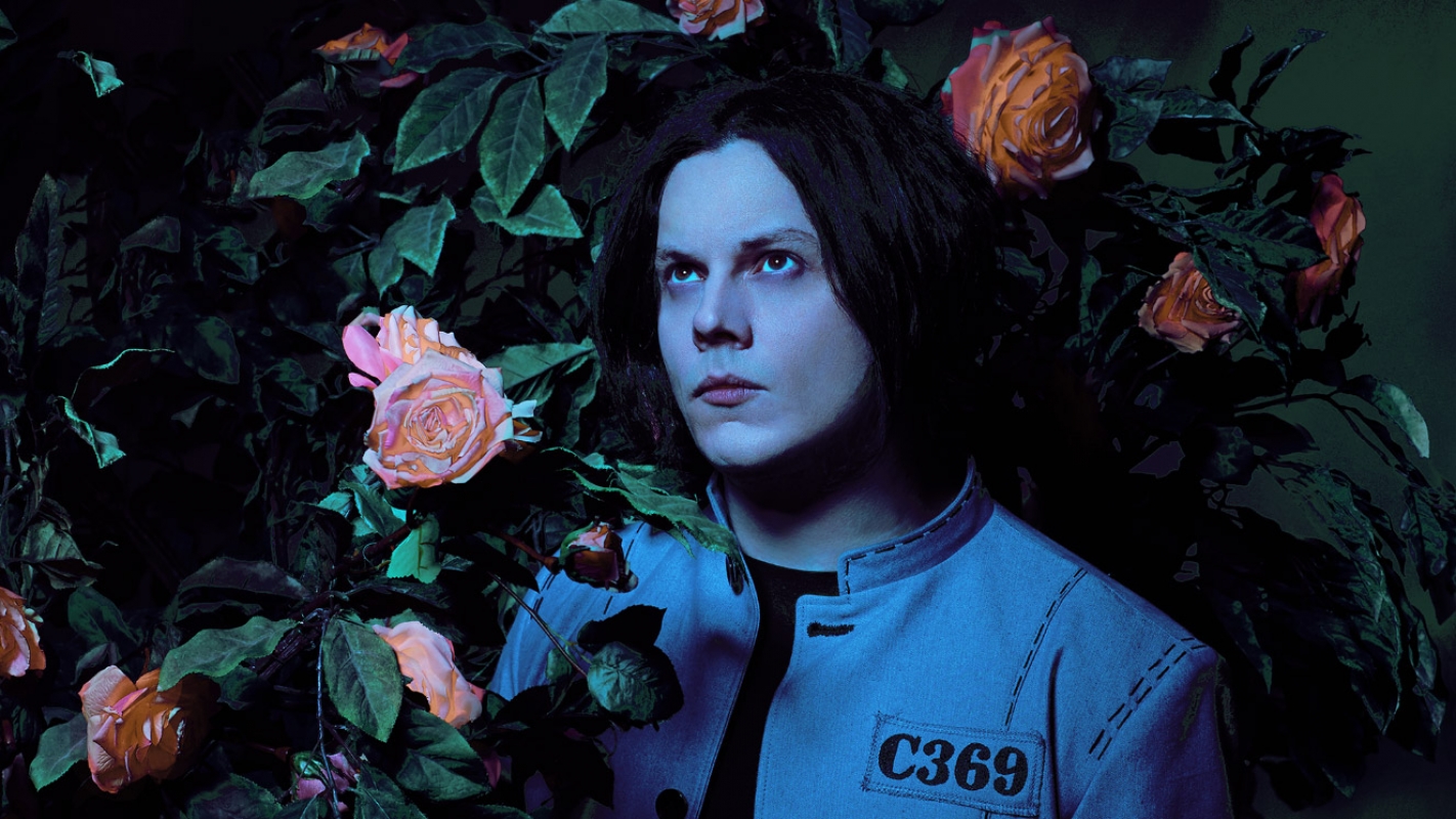 Artistic portrait of a pale-skinned adult male with black hair surrounded by peach roses and green foliage in a dramatic dark lighting.