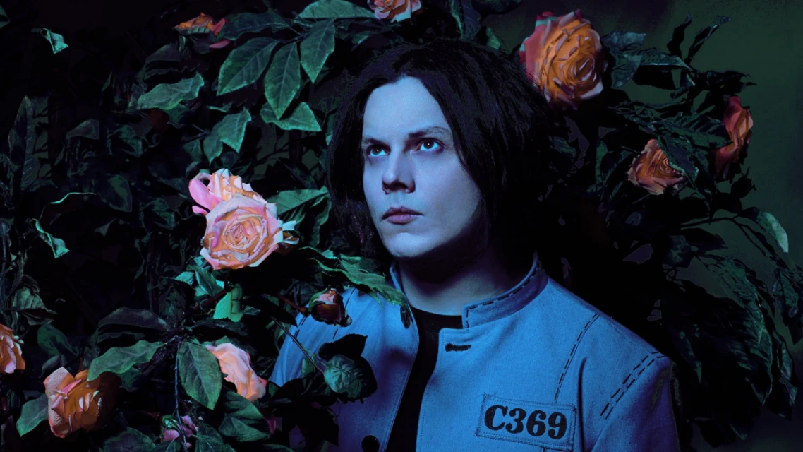 Artistic portrait of a pale-skinned adult male with black hair surrounded by peach roses and green foliage in a dramatic dark lighting.