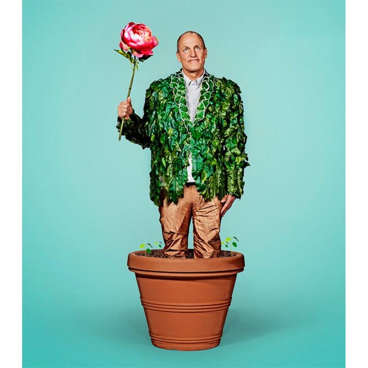 Man standing in a terracotta pot wearing a green leaf jacket and holding a pink flower against a bright teal background.