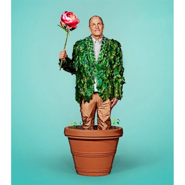 Man standing in a terracotta pot wearing a green leaf jacket and holding a pink flower against a bright teal background.