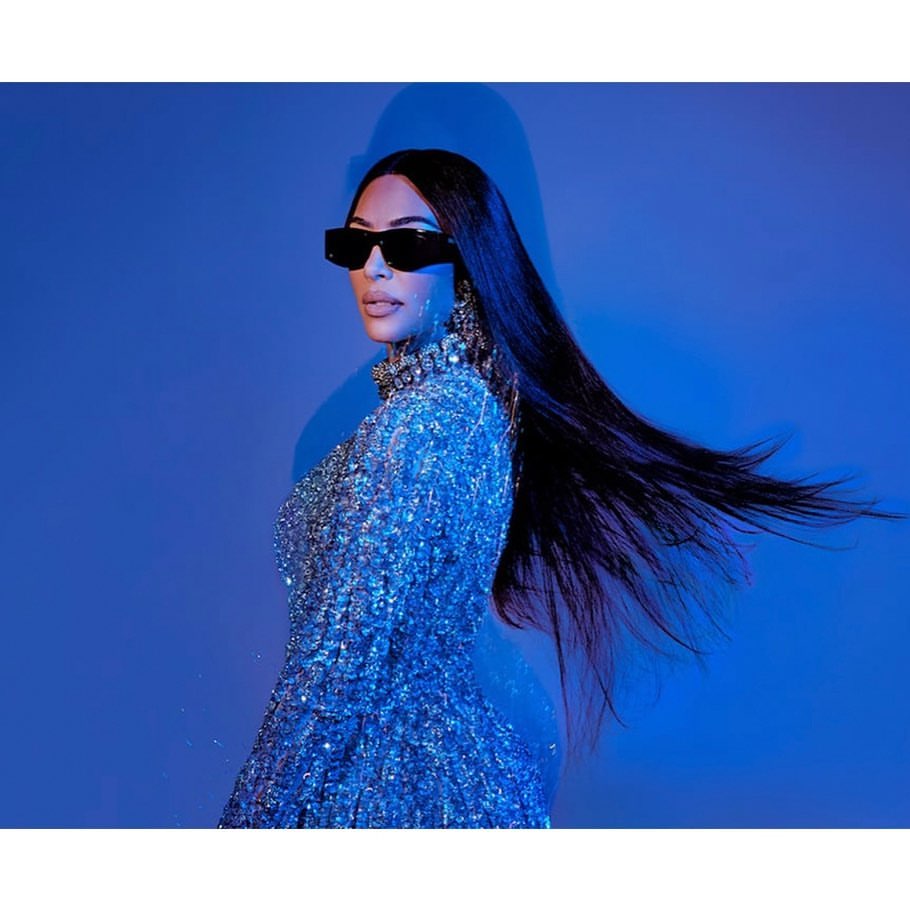 Elegant woman with long black hair poses in a sparkling blue dress and black sunglasses against a blue backdrop, side view.