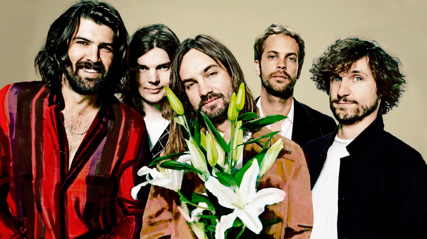 A group of five casually dressed men pose indoors holding a bouquet of white lilies in a medium shot with a neutral background.