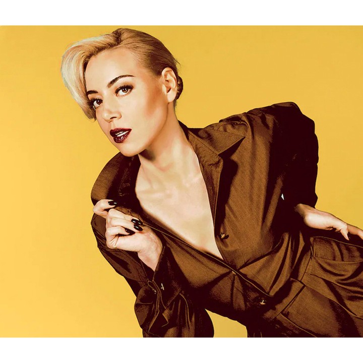 Aubrey Plaza poses in a brown jumpsuit against a yellow backdrop in a studio portrait featuring a close-up shot and eye-level angle.