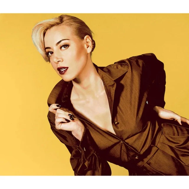 Aubrey Plaza poses in a brown jumpsuit against a yellow backdrop in a studio portrait featuring a close-up shot and eye-level angle.